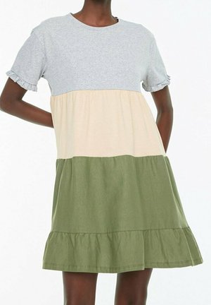 Vrouw die een korte mouwen jurk met kleurvlakken draagt met lichtgrijze, beige en olijfgroene lagen en gerimpelde mouwzomen.