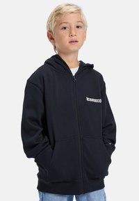 Blond dieťa oblečené v čiernej zapínacej mikine s kapucňou, s rukami v vreckách a svetlomodrými džínsami pred jednoduchým bielym pozadím.