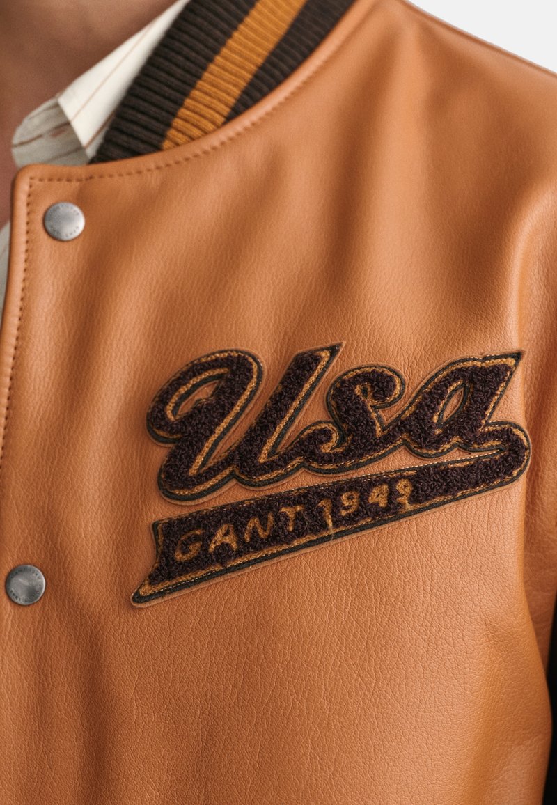 Braune Leder-Baseballjacke mit einem strukturierten, dunkelbraun gestickten "USA"-Logo und "GANT 1948" darunter, akzentuiert durch einen gestreiften Kragen.