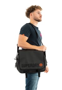 Schwarze Umhängetasche mit "SUPERDRY"-Logo in Orange, aus strapazierfähigem Material gefertigt. Verfügt über einen verstellbaren Schultergurt und eine vordere Klappe mit Schnallen.