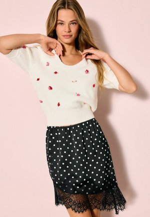 White knitted sweater with floral embroidery, paired with a black polka dot skirt featuring a lace hem. The outfit includes short sleeves.