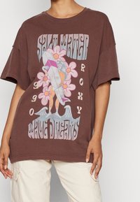 T-shirt marrón de gran tamaño hecho de algodón. Presenta un diseño gráfico con una tabla de surf, flores y texto en varios colores. Mangas cortas, ajuste relajado.
