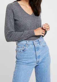 Vrouw in een aansluitende grijze V-halstrui en hoog-geknipte lichtblauwe jeans, met haar handen lichtjes samengevouwen op haar taille tegen een witte achtergrond.