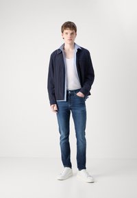 Camisa ligera de marina con un collar azul claro, camiseta blanca y jeans azules oscuros. Los zapatos son blancos, y el conjunto tiene un ajuste relajado.