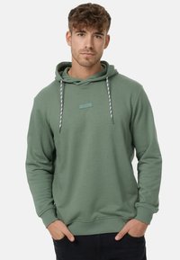Grüne Sweatjacke mit Kapuze, ausgestattet mit einem kleinen Markenetikett, einer Kordel mit schwarz-weißem Muster und gerippten Bündchen und Saum.