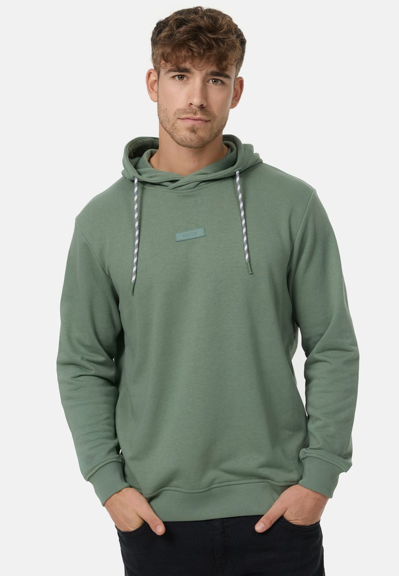 Grüne Sweatjacke mit Kapuze, ausgestattet mit einem kleinen Markenetikett, einer Kordel mit schwarz-weißem Muster und gerippten Bündchen und Saum.