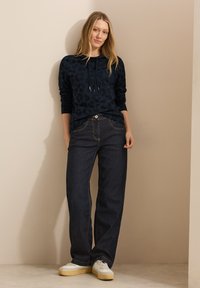 Sweat à capuche en imprimé léopard bleu marine avec cordons, associé à un jean large bleu foncé et à des baskets blanches à semelles jaunes. Arrière-plan texturé simple.