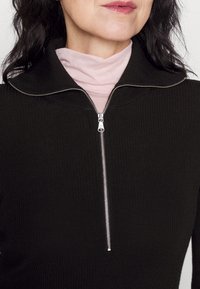 Svart ribbad tröja med hög krage, med en silver dragkedja och en ljusrosa polo under. Mjuk textur och figurnära design.