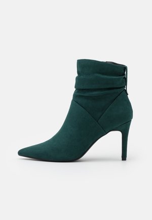 Bottine en daim vert foncé avec un bout pointu, une tige affaissée et un talon haut et fin sur fond blanc.