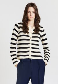 Black and cream striped cardigan with a round neckline, front buttons, and long sleeves, paired with navy trousers. Textured knit material.