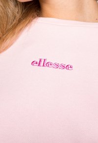 Camiseta de algodón de color rosa claro con un logo bordado en un tono más oscuro de rosa en la parte izquierda del pecho, ligeramente levantado. Textura suave.