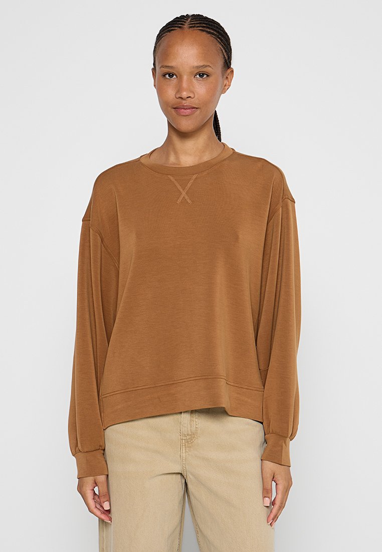 Jong persoon met gevecht haar die een losse bruine sweatshirt en beige broek draagt, staande tegen een effen lichte achtergrond.