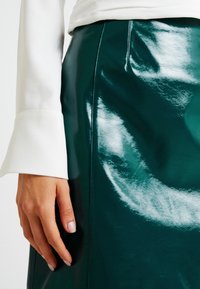 Groene glanzende faux leren rok met een nauwsluitende vorm, voorzien van zichtbare stiksels en een gladde, reflecterende oppervlakte. Witte mouw gedeeltelijk zichtbaar.