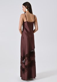 Frau mit langen dunklen Haaren trägt ein ärmelloses braunes Satin-Kleid mit Spitzenpanelen und großen runden Ohrringen, steht vor einem schlichten hellen Hintergrund.