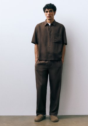 Jeune homme aux cheveux bouclés et foncés, portant une chemise à manches courtes marron à boutons assortie à un pantalon, debout les mains dans les poches contre un mur blanc.