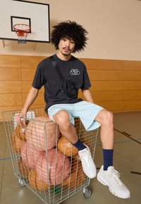Camiseta gráfica negra, pantalones cortos azul claro y zapatillas blancas. Sentado en un carrito de alambre lleno de balones de baloncesto de varios colores en un gimnasio.