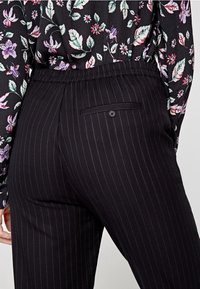 Pantaloni neri a righe con vita elasticizzata. Presentano una tasca posteriore con dettaglio a bottone. Il tessuto ha una texture liscia.