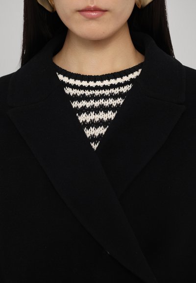 Schwarzer Wollmantel mit Reverskragen über einem gestreiften Strickpullover in Schwarz und Creme, mit einem gerippten Halsauschnitt. Glatte Textur.