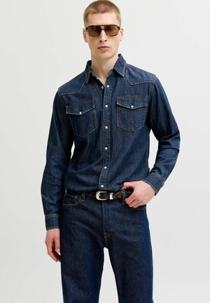 Jeune homme portant une chemise en denim foncé et un jean avec une ceinture tressée noire, posant avec une main sur la boucle de la ceinture et portant des lunettes de soleil.