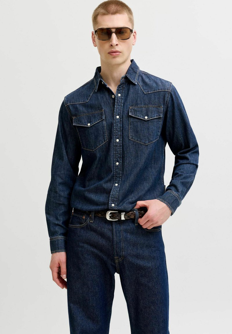 Jeune homme portant une chemise en denim foncé et un jean avec une ceinture tressée noire, posant avec une main sur la boucle de la ceinture et portant des lunettes de soleil.