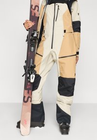 Beige és fekete síruha vízálló anyagból, cipzáras zsebekkel és megerősített térdekkel. Síszárakat tartó személy fekete kötésekkel.