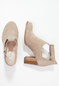 Beige mockaskinnsstövlar vid ankeln med öppna sidor, rund tå och en texturerad träklack. Har en silverfärgad spänne på sidan.