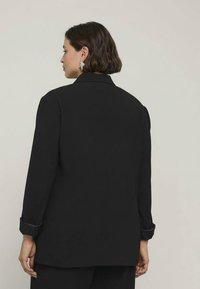 Černý blejzr s dlouhými rukávy a volným střihem. Obsahuje klopový límec a ohrnuté manžety. Hladká látka s mírným leskem.