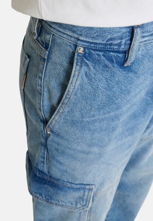 Primer plano de pantalones cargo de mezclilla azul con bolsillos laterales y costuras visibles, combinados con una camiseta blanca.