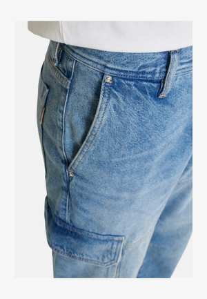 Primer plano de pantalones cargo de mezclilla azul con bolsillos laterales y costuras visibles, combinados con una camiseta blanca.