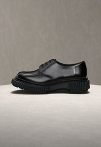 Chaussure habillée en cuir noir avec une semelle en caoutchouc épaisse et rainurée et trois œillets pour les lacets, présentée sur une surface claire avec un fond gris.