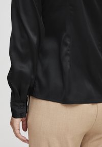 Schwarzes Satin-Hemd mit langen Ärmeln und abgerundetem Saum. Verfügt über geknöpfte Manschetten und eine glatte Textur, kombiniert mit hellbeigen maßgeschneiderten Hosen.