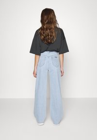 Jeans de perna larga azul clara com cintura alta, apresentando bolsos traseiros em patch e uma textura de denim suave. Combinados com uma camisa solta escura.