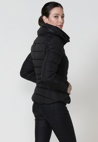 Chaqueta de abrigo negra con cuello alto, diseño acolchado y silueta ajustada. Textura suave y bolsillos con cremallera visibles.