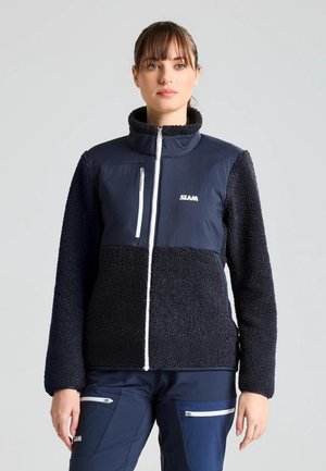 Una mujer luce una chaqueta de forro polar azul marino y negra, con cremalleras blancas y pantalones a juego, sobre un fondo gris liso.