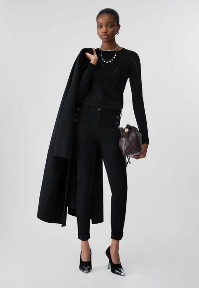 Femme portant un pull et un pantalon noirs, tenant un manteau noir sur une épaule et un sac marron foncé dans une main.