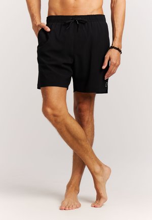 Man in zwarte zwemshorts met elastische tailleband en koordje, staand op blote voeten met één been gekruist voor het andere.