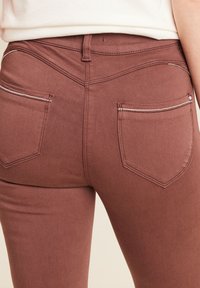Bruine, rekbare broek met een gladde textuur, voorzien van twee achterzakken en contrasterende stiksels langs de zakken voor extra detail.