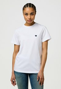Hvid bomulds T-shirt med korte ærmer, rund halsudskæring og et lille marineblåt logo på venstre bryst. Sammen med tætsiddende denim jeans.