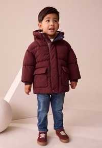 Veste matelassée bordeaux avec capuche, dotée d'une fermeture éclair sur le devant, de deux poches avant et d'une doublure intérieure chaude, associée à un jean bleu et des baskets bordeaux.