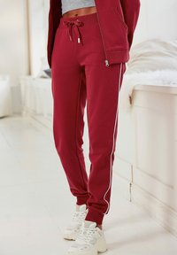 Burgunderfarbene Sweatpants aus weichem Stoff, mit einem Kordelzugbund, seitlichen weißen Streifen und engen Abschlüssen, kombiniert mit weißen Sneakers.