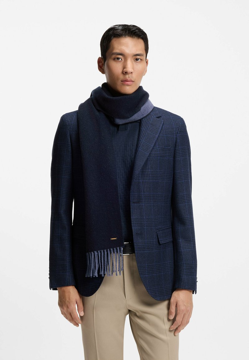 Mørkeblå halsklæde lavet af blødt stof, med en mørkere nuance og frynser i enderne, båret over en struktureret, mørk sweater og mønstret blazer.