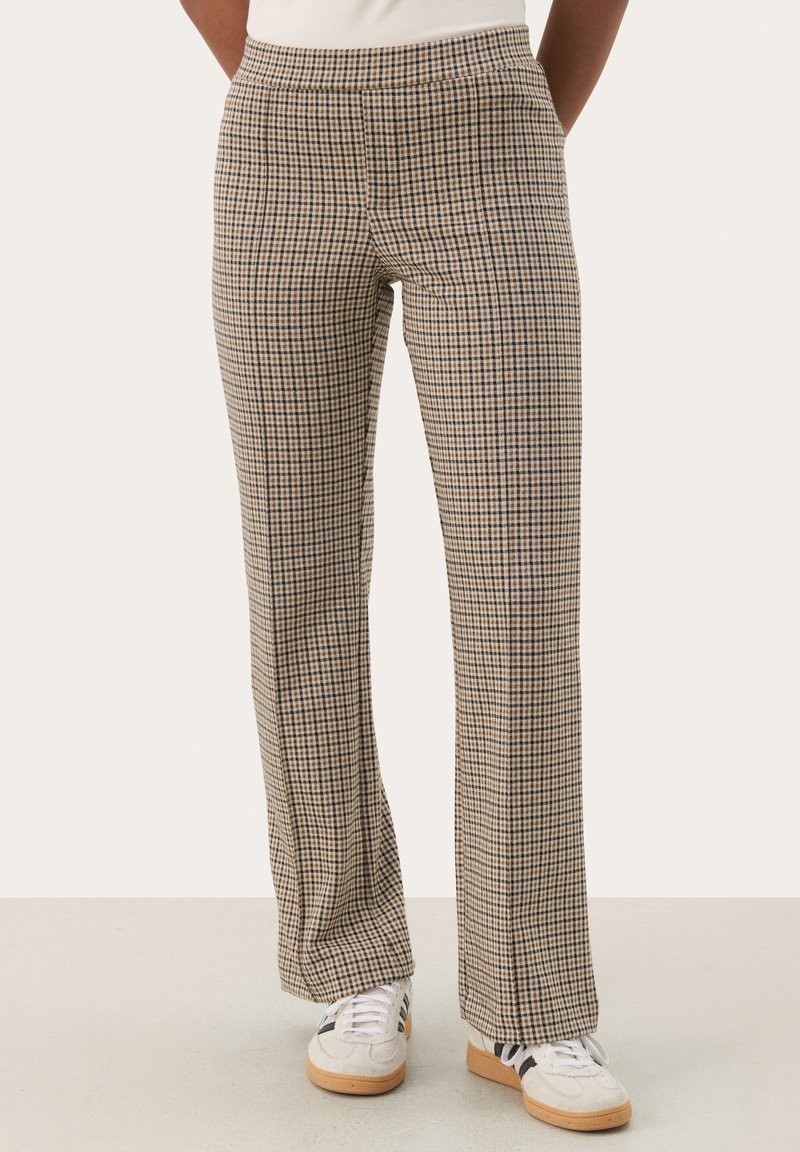 Geblokte broek met een beige en bruine print, voorzien van een losse pasvorm, elastische tailleband en subtiele textuur. Gekleed met witte sneakers.