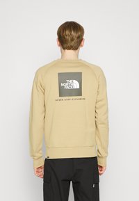 Sudadera beige con cuello redondo y mangas raglán. Presenta un logo cuadrado en verde oscuro con texto blanco en la parte posterior.