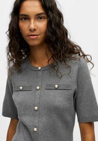 Pull en tricot gris à manches courtes avec des boutons en forme de perles, deux poches poitrine et un col rond avec des accents côtelés.