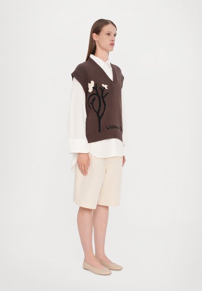 Brauner Strickwesten mit V-Ausschnitt, verziert mit floralen Mustern in Creme und Schwarz. Getragen über einem weißen Hemd und kombiniert mit beigen Shorts.