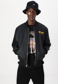 Giubbotto bomber nero con zip frontale, colletto in maglia e logo giallo "Koltak". Indossato sopra una t-shirt grafica nera e abbinato a pantaloni neri.