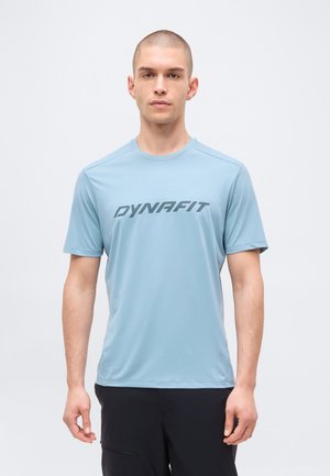 Νέος άνδρας με ξυρισμένο κεφάλι, που φοράει ένα ανοιχτό μπλε κοντομάνικο Dynafit μπλουζάκι και μαύρο παντελόνι, στέκεται μπροστά σε μονόχρωμο φόντο.