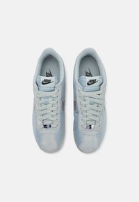 Világoskék Nike sportcipők, amelyek bőrből és velúrból készültek, párnázott nyelvvel, texturált felszínnel és szürke fűzőkkel rendelkeznek, fekete logóval.