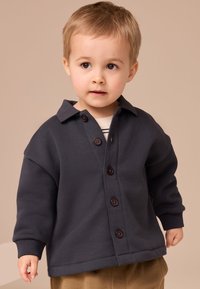 Sötétszürke gombos kabát gallérral, barna gombokkal és bő ujjakkal, fehér csíkos aláöltözővel párosítva.