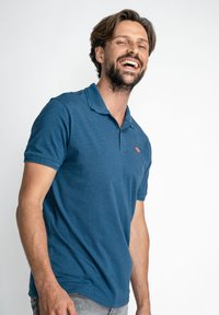 Camisa polo de rayas azules con cuello, que presenta un pequeño logo rojo en el lado izquierdo del pecho. La tela parece suave y tiene un ajuste casual.